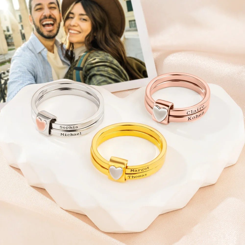 Three Anillos personalizados con nombres-uno en plata, uno en oro y uno en oro rosa de acero inoxidable con un pequeño corazón-se exhiben sobre un plato en forma de corazón; al fondo se ve la foto sonriente de una pareja.