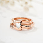 Tres Anillos personalizados con nombres en oro rosa se apilan juntos, sostenidos por un charm de plata en forma de corazón. Cada anillo lleva grabados los nombres de Ryan, Emily y Jacob. El fondo está suavemente difuminado.