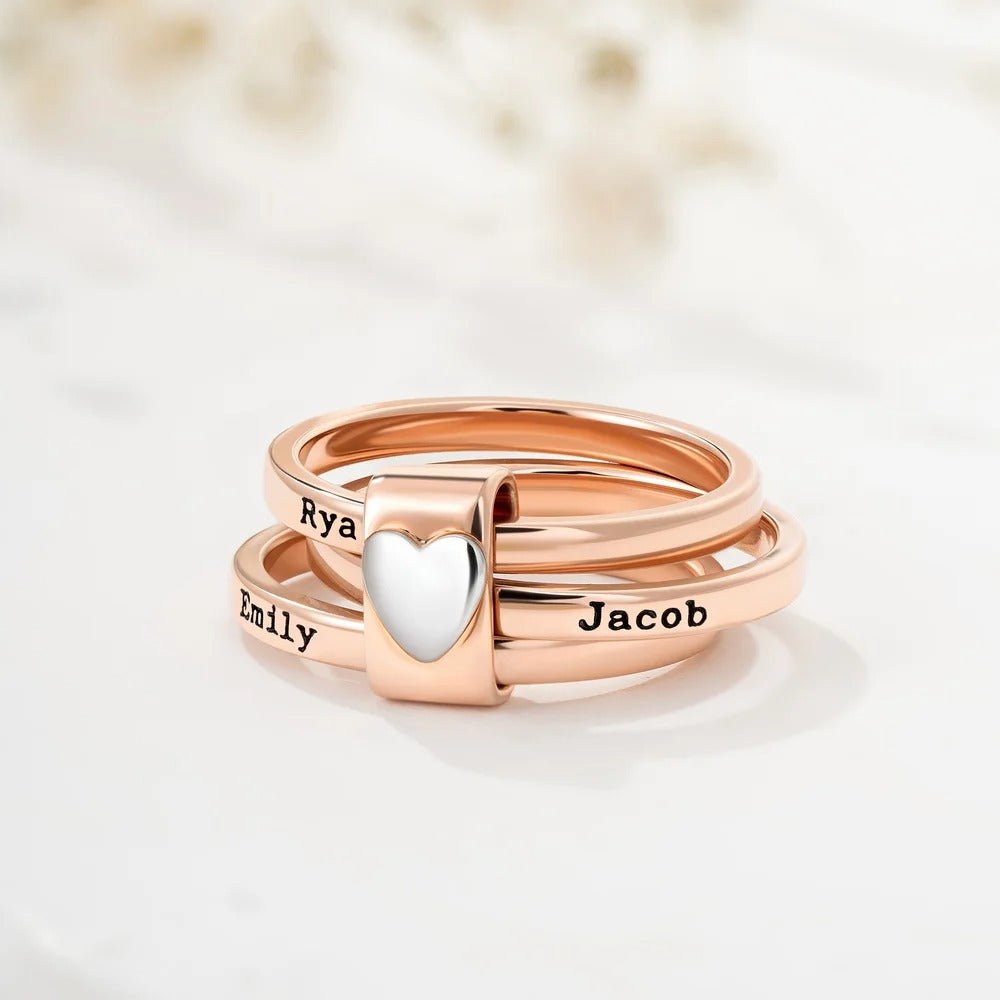 Tres Anillos personalizados con nombres en oro rosa se apilan juntos, sostenidos por un charm de plata en forma de corazón. Cada anillo lleva grabados los nombres de Ryan, Emily y Jacob. El fondo está suavemente difuminado.