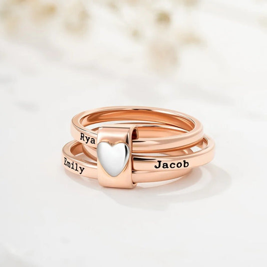 Tres Anillos personalizados con nombres en oro rosa se apilan juntos, sostenidos por un charm de plata en forma de corazón. Cada anillo lleva grabados los nombres de Ryan, Emily y Jacob. El fondo está suavemente difuminado.