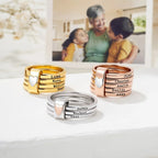 Anillos personalizados con nombres: tres anillos apilables en oro, oro rosa y plata de acero inoxidable, cada uno grabado con nombres y decorado con un corazón. Exhibidos sobre una superficie blanca con una foto borrosa de fondo.