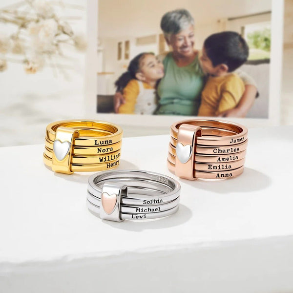 Anillos personalizados con nombres: tres anillos apilables en oro, oro rosa y plata de acero inoxidable, cada uno grabado con nombres y decorado con un corazón. Exhibidos sobre una superficie blanca con una foto borrosa de fondo.