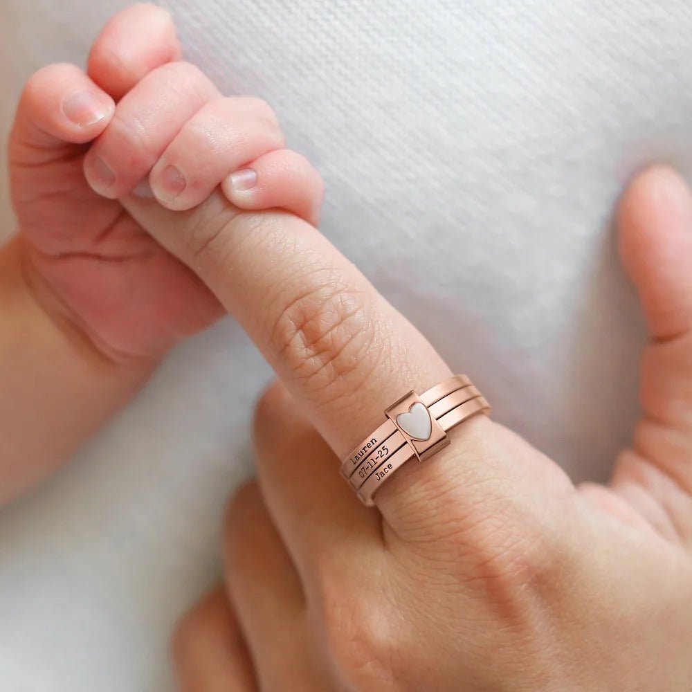 Una mano adulta que lleva un Anillo personalizado con nombres y detalle de corazón sostiene suavemente la mano de un bebé, que agarra el dedo del adulto sobre un suave fondo blanco.