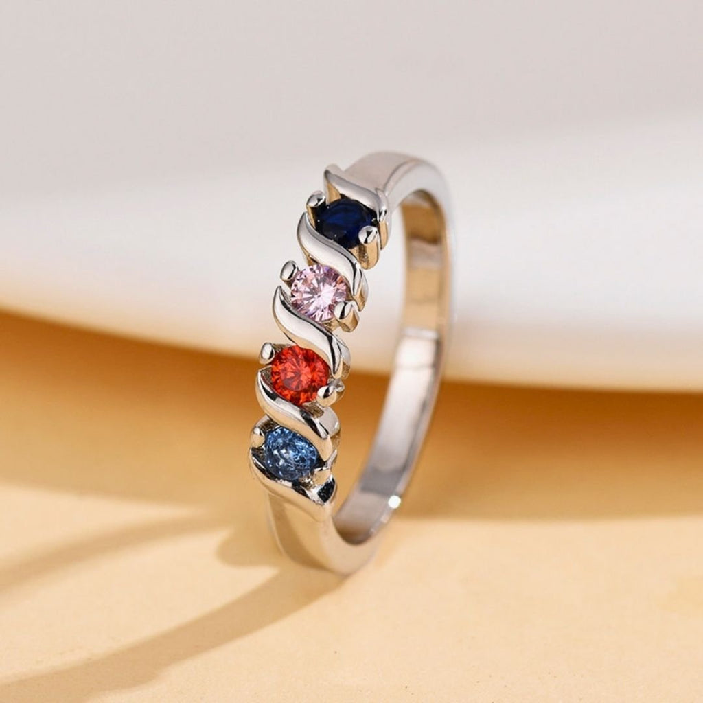 El Anillo personalizado con piedras familiares, color plateado, luce cuatro gemas azul oscuro, rosa claro, rojo y verde azulado en un moderno diseño ondulado de garras; ideal como anillo de piedras de nacimiento sobre fondo beige.