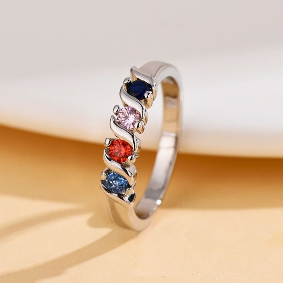 El Anillo personalizado con piedras familiares, color plateado, luce cuatro gemas azul oscuro, rosa claro, rojo y verde azulado en un moderno diseño ondulado de garras; ideal como anillo de piedras de nacimiento sobre fondo beige.