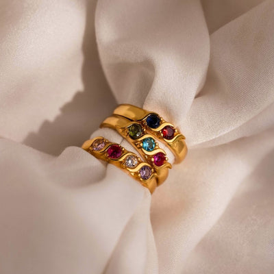 Tres anillos dorados "Anillo personalizado con piedras familiares" lucen gemas en rojo, azul, verde y púrpura sobre tela marfil, apilados para un look único y personalizado.