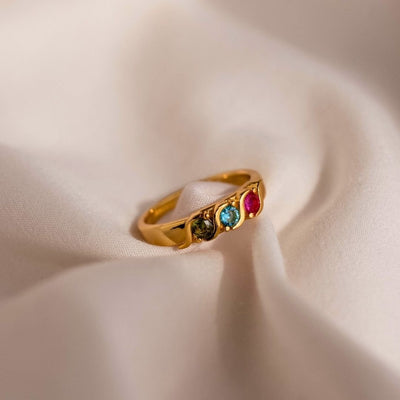Anillo personalizado con piedras familiares en oro, con tres gemas-verde, azul y rojo-alineadas, exhibido sobre suave tela beige clara.