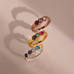 Tres Anillos personalizados con piedras familiares en oro rosa, oro amarillo y plata -cada uno engastado con cinco piedras preciosas de colores- están dispuestos verticalmente, proyectando suaves sombras sobre una superficie de color beige claro.