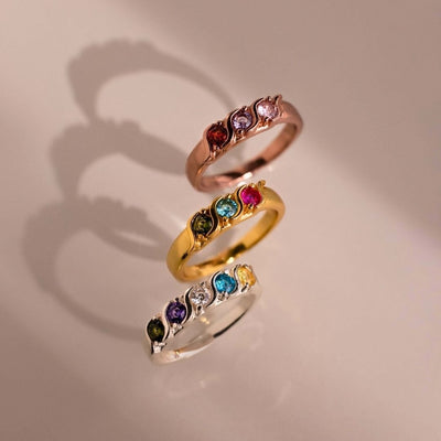 Tres Anillos personalizados con piedras familiares en oro rosa, oro amarillo y plata -cada uno engastado con cinco piedras preciosas de colores- están dispuestos verticalmente, proyectando suaves sombras sobre una superficie de color beige claro.