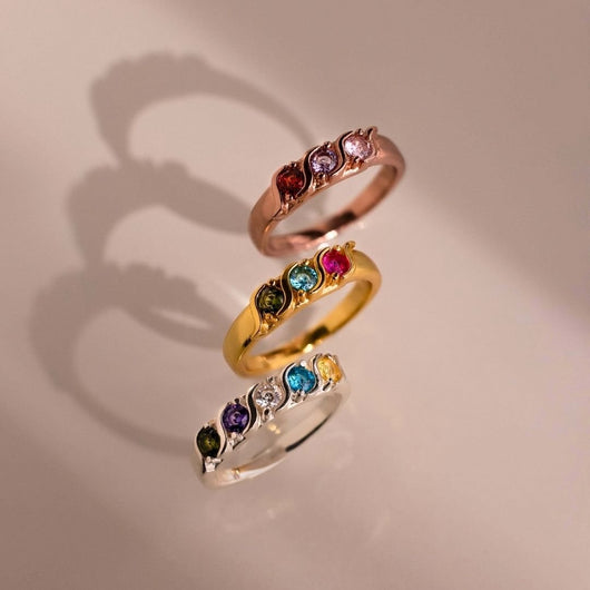 Tres Anillos personalizados con piedras familiares en oro rosa, oro amarillo y plata -cada uno engastado con cinco piedras preciosas de colores- están dispuestos verticalmente, proyectando suaves sombras sobre una superficie de color beige claro.
