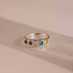 Anillo personalizado con piedras familiares: este anillo plateado presenta cinco gemas redondas en verde, morado, transparente, azul y amarillo alineadas sobre la banda. Se muestra sobre una superficie lisa y clara.