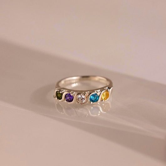 Anillo personalizado con piedras familiares: este anillo plateado presenta cinco gemas redondas en verde, morado, transparente, azul y amarillo alineadas sobre la banda. Se muestra sobre una superficie lisa y clara.