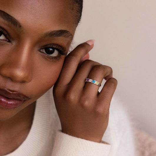Una mujer de piel morena suave y maquillaje natural mira suavemente a la cámara, acercándose la mano a la cara para mostrar un Anillo personalizado con piedras familiares. Lleva un jersey de color crema claro.