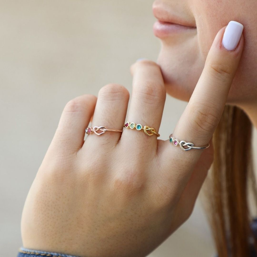 Un primer plano de la mano de una mujer de piel clara, cuidada con esmalte de uñas blanco, muestra tres delicados Anillo personalizado Lazos del corazón anillos adornados con pequeñas piedras de nacimiento de colores en sus dedos.