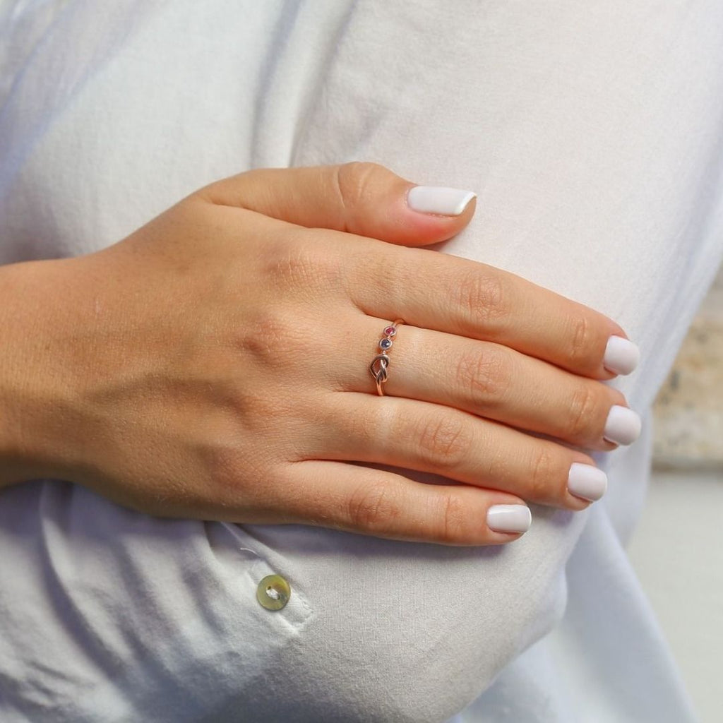 Una mano con las uñas cuidadas y de color rosa pálido descansa sobre la manga de una camisa blanca, mostrando el Anillo personalizado Lazos del corazón adornado con piedras de nacimiento en el dedo anular.