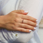 Una mano con las uñas cuidadas y de color rosa pálido descansa sobre la manga de una camisa blanca, mostrando el Anillo personalizado Lazos del corazón adornado con piedras de nacimiento en el dedo anular.