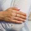 Una mano con las uñas cuidadas y de color rosa pálido descansa sobre la manga de una camisa blanca, mostrando el Anillo personalizado Lazos del corazón adornado con piedras de nacimiento en el dedo anular.