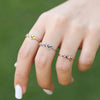 Una mano cuidada luce tres anillos Anillo personalizado Lazos del corazón -oro, plata y oro rosa-, cada uno con una pequeña piedra de nacimiento engastada sobre un fondo verde difuminado.
