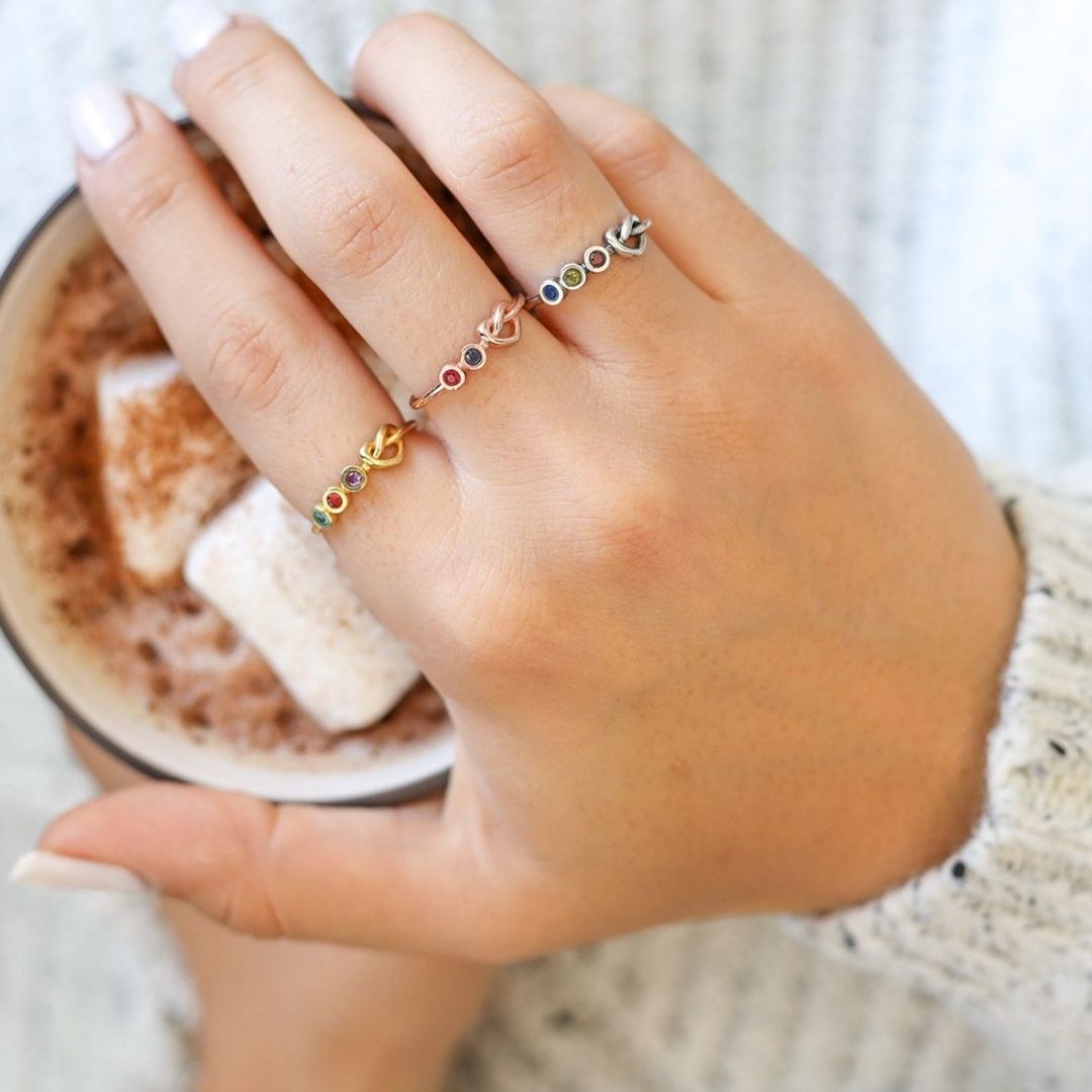 Una mano sostiene una taza de chocolate caliente con malvaviscos y lleva tres anillos Anillo personalizado Lazos del corazón adornados con piedras preciosas de colores, sobre un acogedor fondo de jersey de punto blanco.