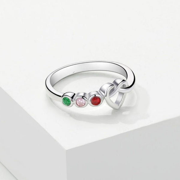 Anillo personalizado Lazos del corazón en color plateado, con tres piedras redondas verdes, rosas y rojas representando piedras de nacimiento y diseño de nudo al lado, expuesto sobre una superficie blanca geométrica.