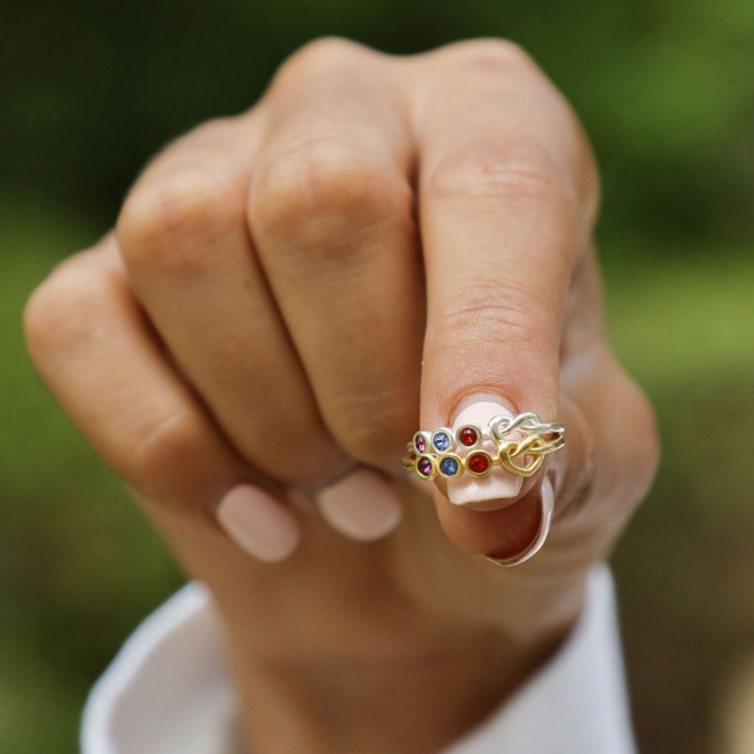 En un fondo verde difuminado aparece una mano con las uñas pintadas de rosa pálido, que lleva el Anillo personalizado Lazos del corazón en el dedo corazón.
