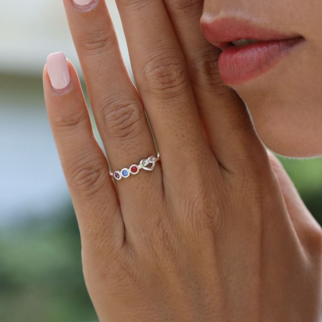 Primer plano de la mano cuidada de una mujer que lleva el Anillo personalizado Lazos del corazón, adornado con piedras de nacimiento de colores, sostenida cerca de su cara con los labios suaves visibles en el fondo.