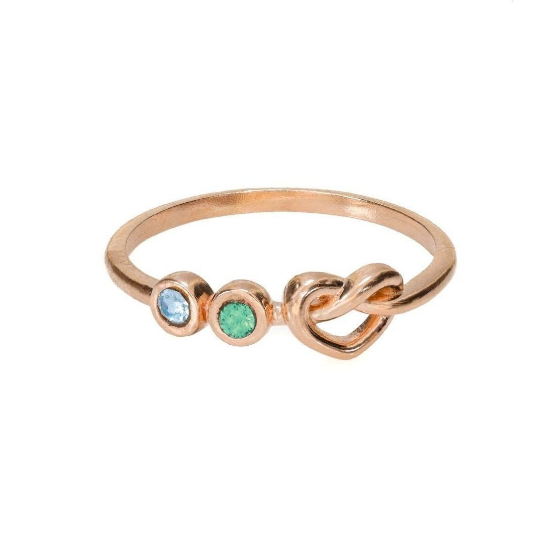 Anillo personalizado Lazos del corazón en oro rosa, con dos piedras de nacimiento azul y verde junto a un diseño de lazo sencillo, fotografiado sobre fondo blanco.