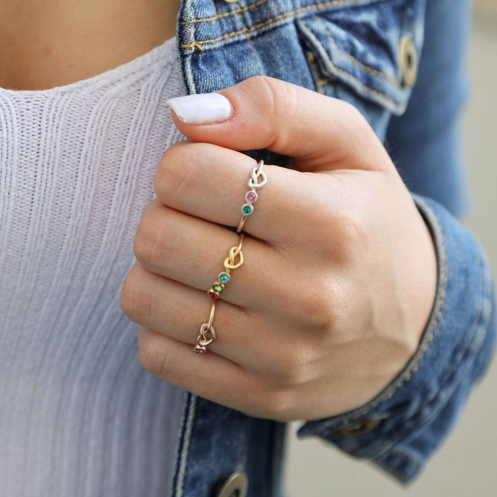 Una mano con manicura blanca lleva dos anillos Anillo personalizado Lazos del corazón con piedras preciosas de colores, sujetando el borde de una cazadora vaquera sobre un top blanco de canalé.