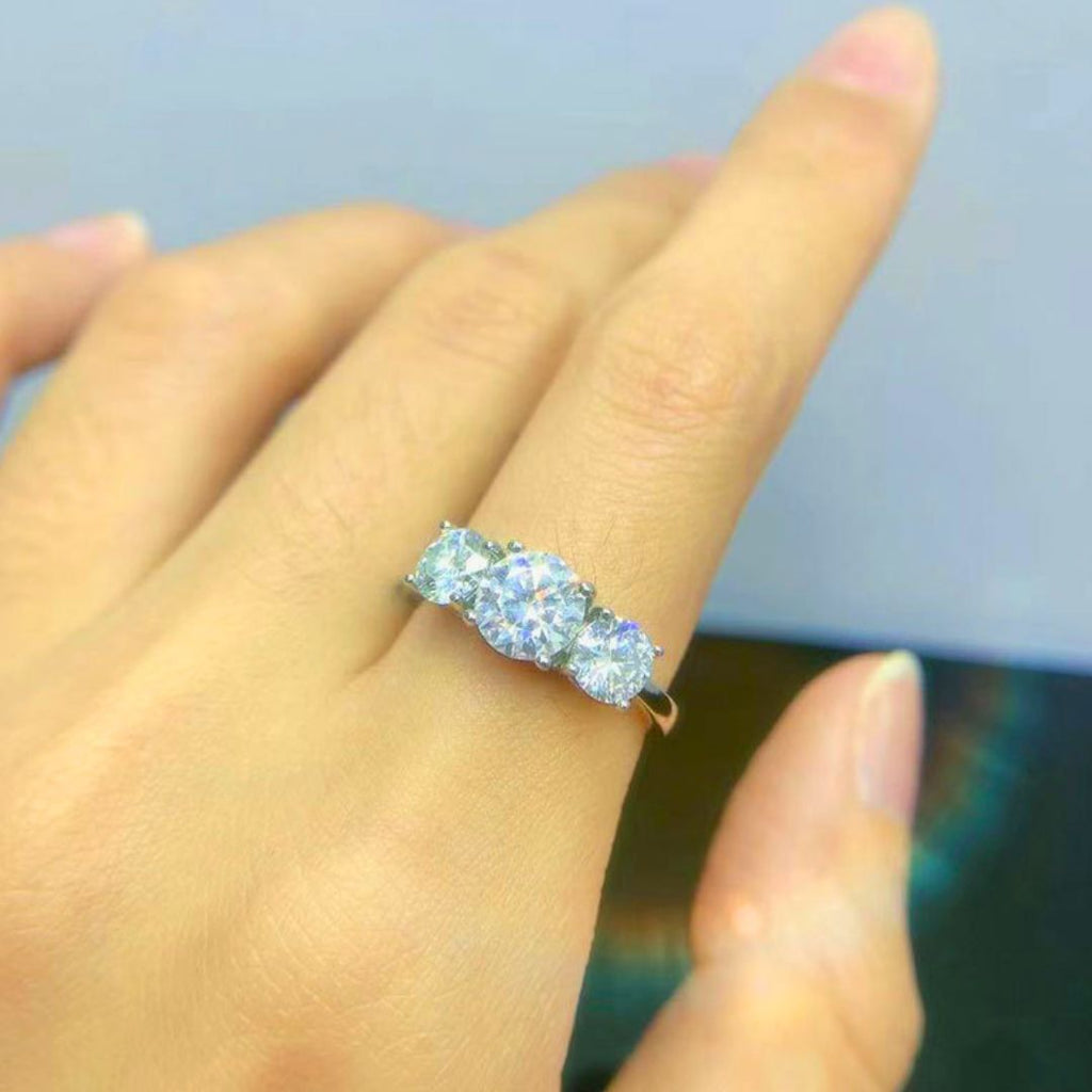 Una mano luce en el dedo anular el Anillo Tres destellos de amor, un anillo de acero inoxidable dorado con tres brillantes diamantes redondos, sobre un fondo suavemente difuminado.