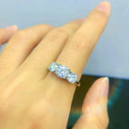 Una mano luce en el dedo anular el Anillo Tres destellos de amor, un anillo de acero inoxidable dorado con tres brillantes diamantes redondos, sobre un fondo suavemente difuminado.