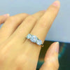 Una mano luce en el dedo anular el Anillo Tres destellos de amor, un anillo de acero inoxidable dorado con tres brillantes diamantes redondos, sobre un fondo suavemente difuminado.