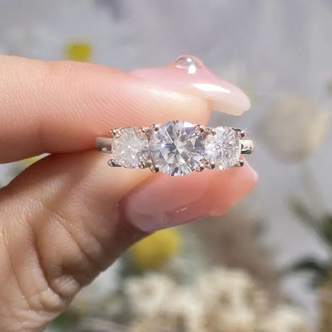 Una mano con las uñas cuidadas sostiene el Anillo Tres destellos de amor, con una gran piedra central flanqueada por dos más pequeñas sobre una elegante banda de plata. El fondo suavemente difuminado añade un toque elegante a la escena.