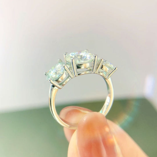 Una mano sostiene el Anillo Tres destellos de amor, un anillo de acero inoxidable con tres brillantes gemas redondas engarzadas en garras, sobre un fondo suavemente difuminado.