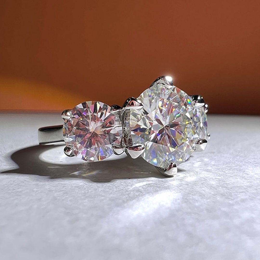 Primer plano del Anillo Tres destellos de amor, elaborado en plata con tres piedras redondas brillantes -una prominente en el centro y dos más pequeñas a cada lado- engastadas sobre un suave fondo naranja y blanco.