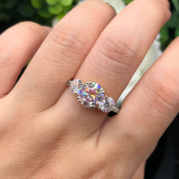 Una mano luce el Anillo Tres destellos de amor, con tres brillantes diamantes talla oval en acero inoxidable. La luz de colores se refleja en el elegante anillo, mientras las hojas verdes se difuminan suavemente en el fondo, resaltando su brillo.