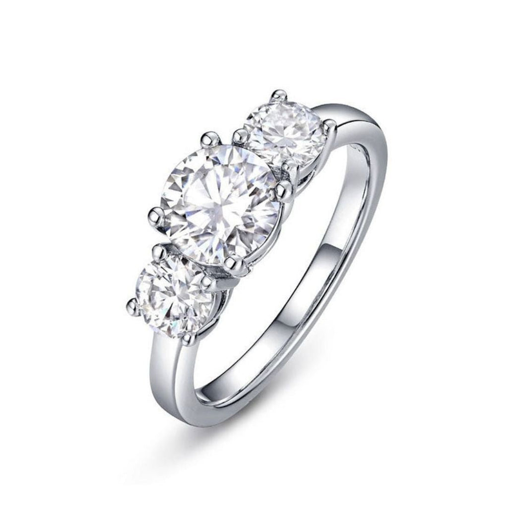 El Anillo Tres destellos de amor es un anillo de acero inoxidable plateado con tres diamantes blancos redondos y brillantes engarzados en fila sobre una banda lisa, que se muestra sobre un fondo blanco.
