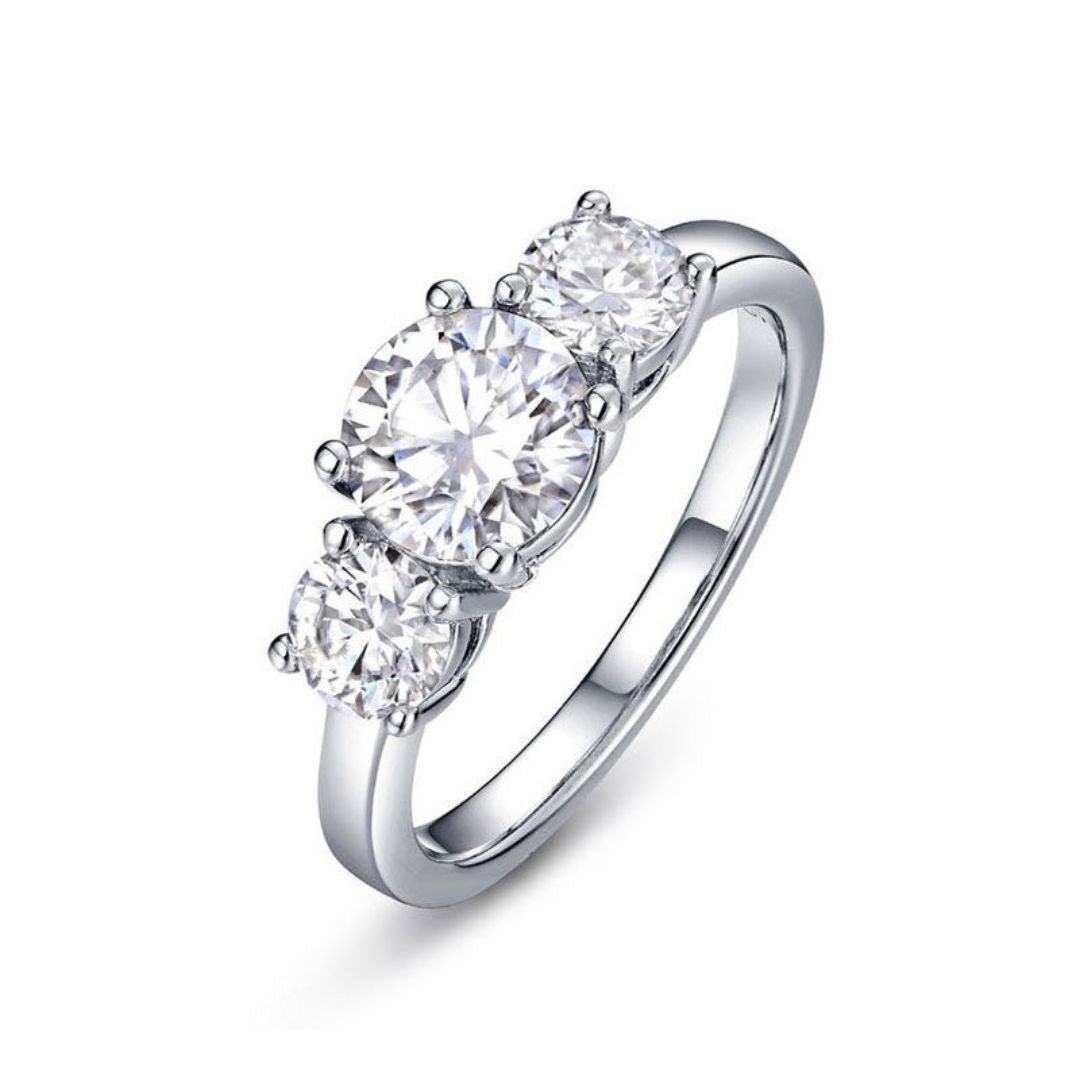 El Anillo Tres destellos de amor es un anillo de acero inoxidable plateado con tres diamantes blancos redondos y brillantes engarzados en fila sobre una banda lisa, que se muestra sobre un fondo blanco.