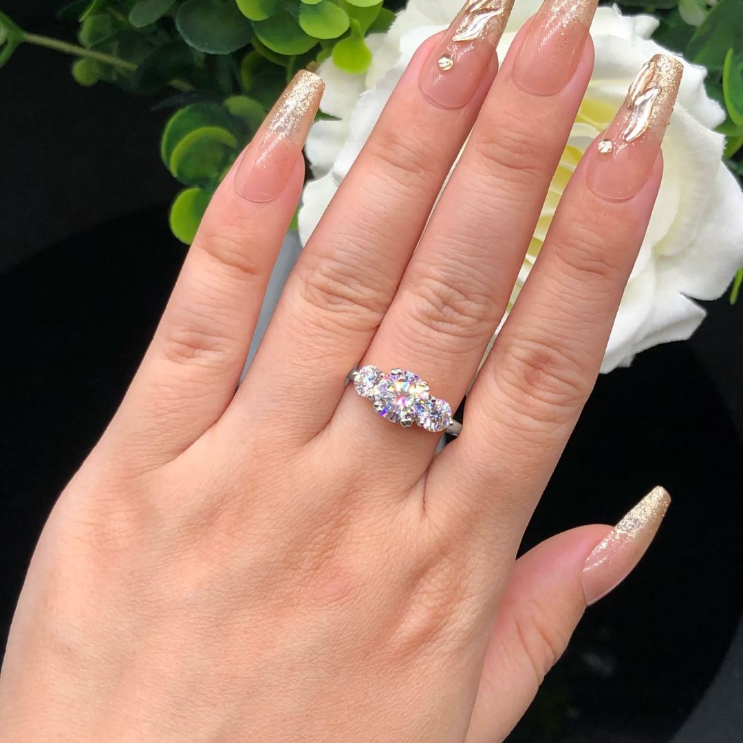 Una mano con uñas largas, doradas y brillantes luce el Anillo Tres destellos de amor en el dedo anular, sus tres piedras centelleantes brillan junto a una flor blanca y hojas verdes de fondo.