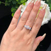 Una mano con uñas largas, doradas y brillantes luce el Anillo Tres destellos de amor en el dedo anular, sus tres piedras centelleantes brillan junto a una flor blanca y hojas verdes de fondo.