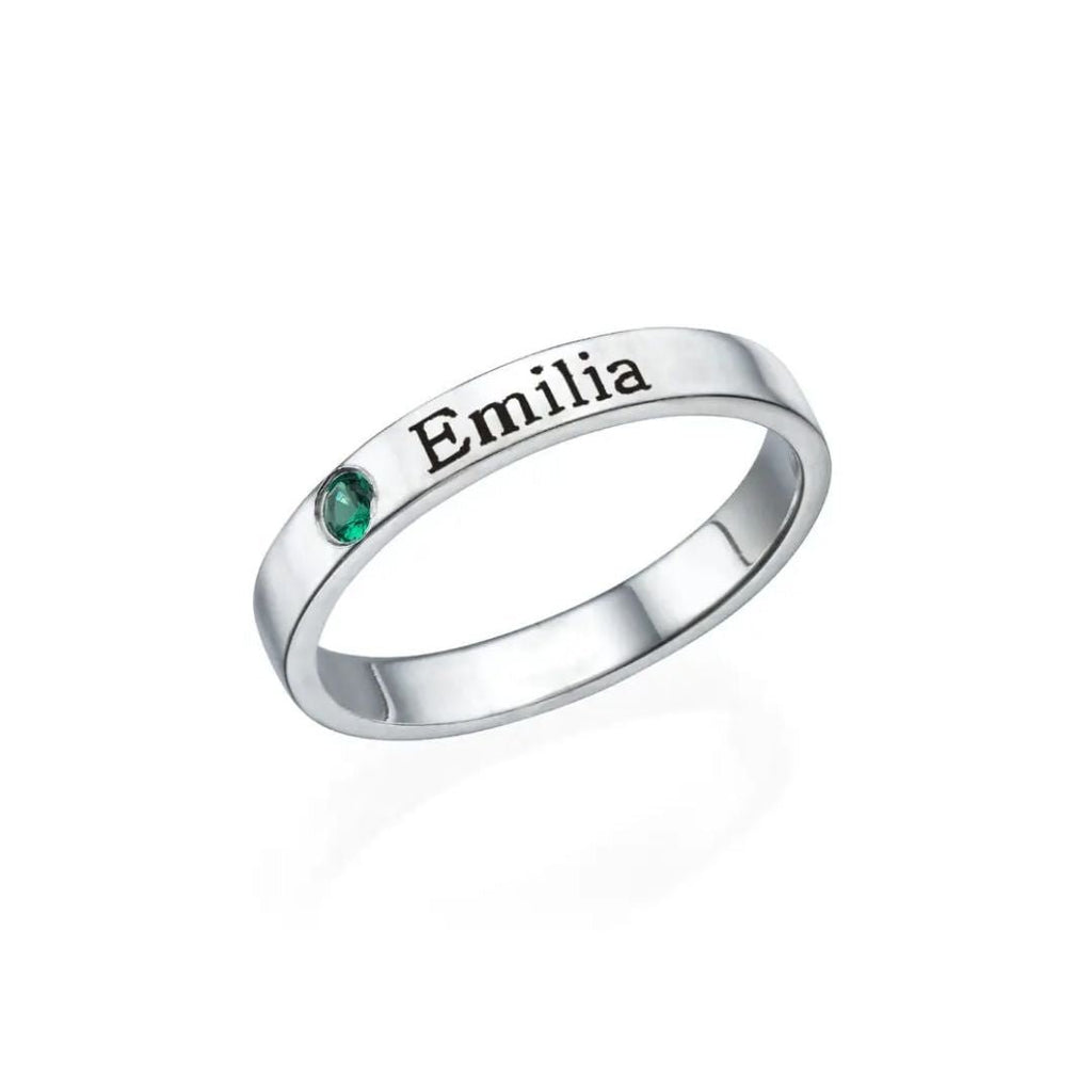 Un Anillos apilables de plata con nombres y piedras de nacimiento, grabado con "Emilia" en negro, lleva una pequeña piedra de nacimiento verde engastada junto al nombre. El anillo personalizado se muestra sobre un fondo blanco liso.