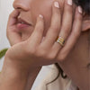 Una mujer apoya la cara en la mano y lleva tres anillos apilables con nombres y piedras de nacimiento. Cada anillo lleva grabados nombres y piedras preciosas. Lleva las uñas pintadas de rosa claro.