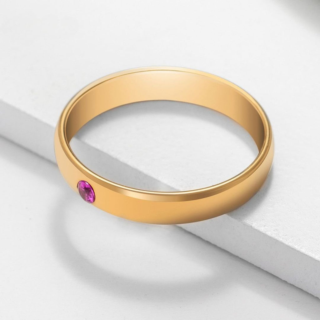 El anillo dorado "Anillos apilables con nombres y piedras de nacimiento" luce una piedra rosa redonda engastada al ras, mostrada sobre una superficie blanca de líneas limpias y modernas.