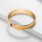 El anillo dorado "Anillos apilables con nombres y piedras de nacimiento" luce una piedra rosa redonda engastada al ras, mostrada sobre una superficie blanca de líneas limpias y modernas.