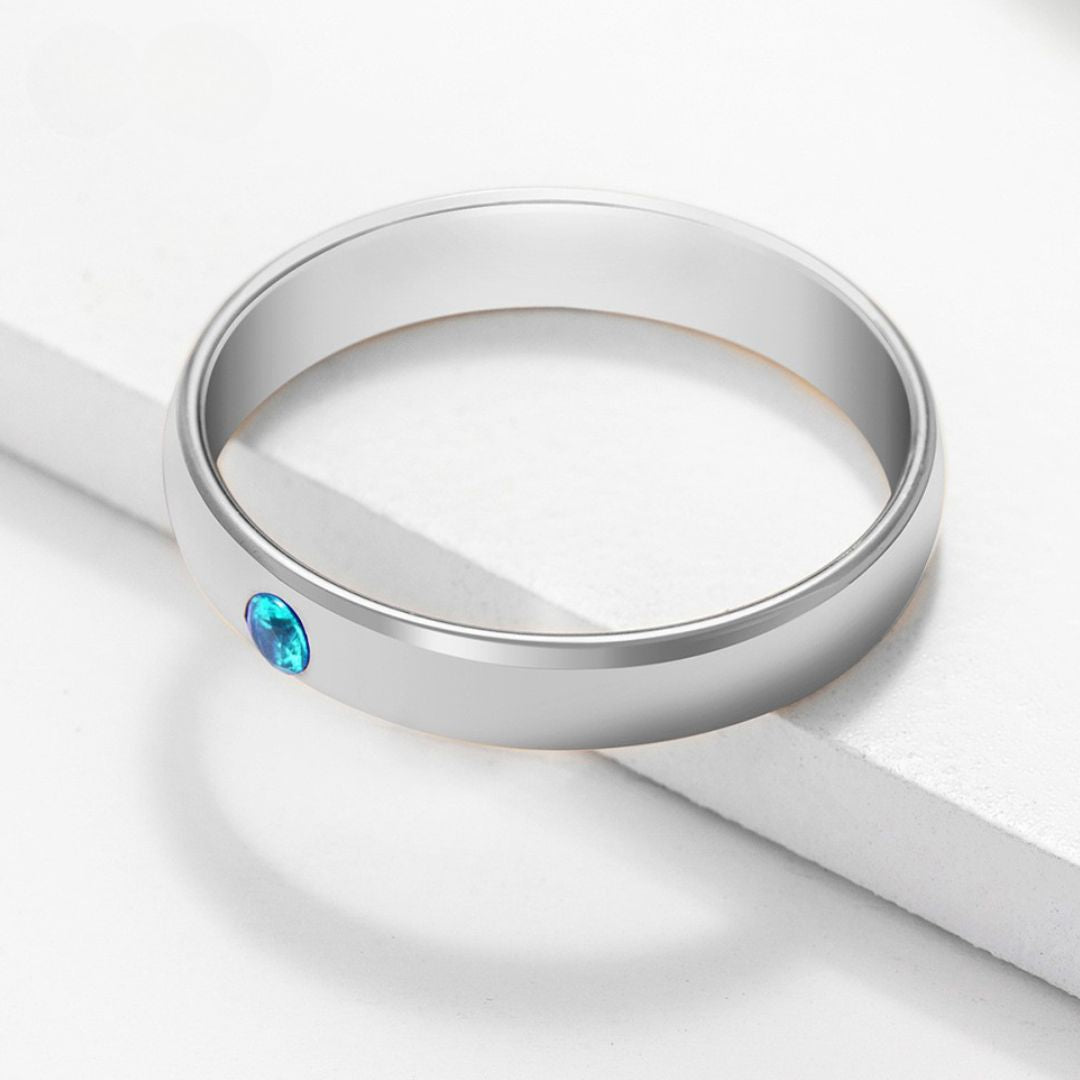 A simple Anillos apilables con nombres y piedras de nacimiento en color plateado, con una pequeña gema azul redonda incrustada en la banda, exhibidos sobre una superficie blanca bajo luz suave.