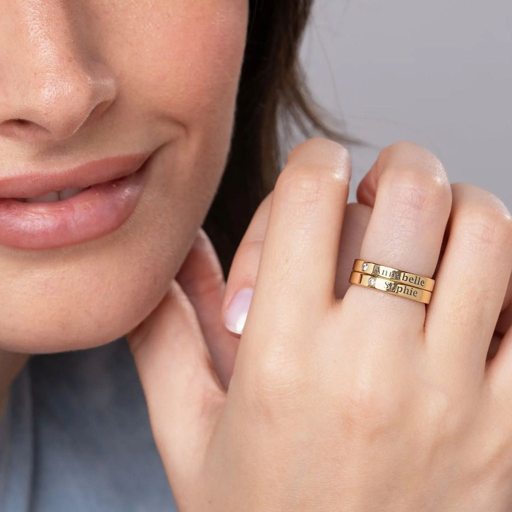 Una mujer sonríe suavemente, mostrando su mano adornada con dos Anillos apilables con nombres y piedras de nacimiento grabados con "ma belle" y "sophie".