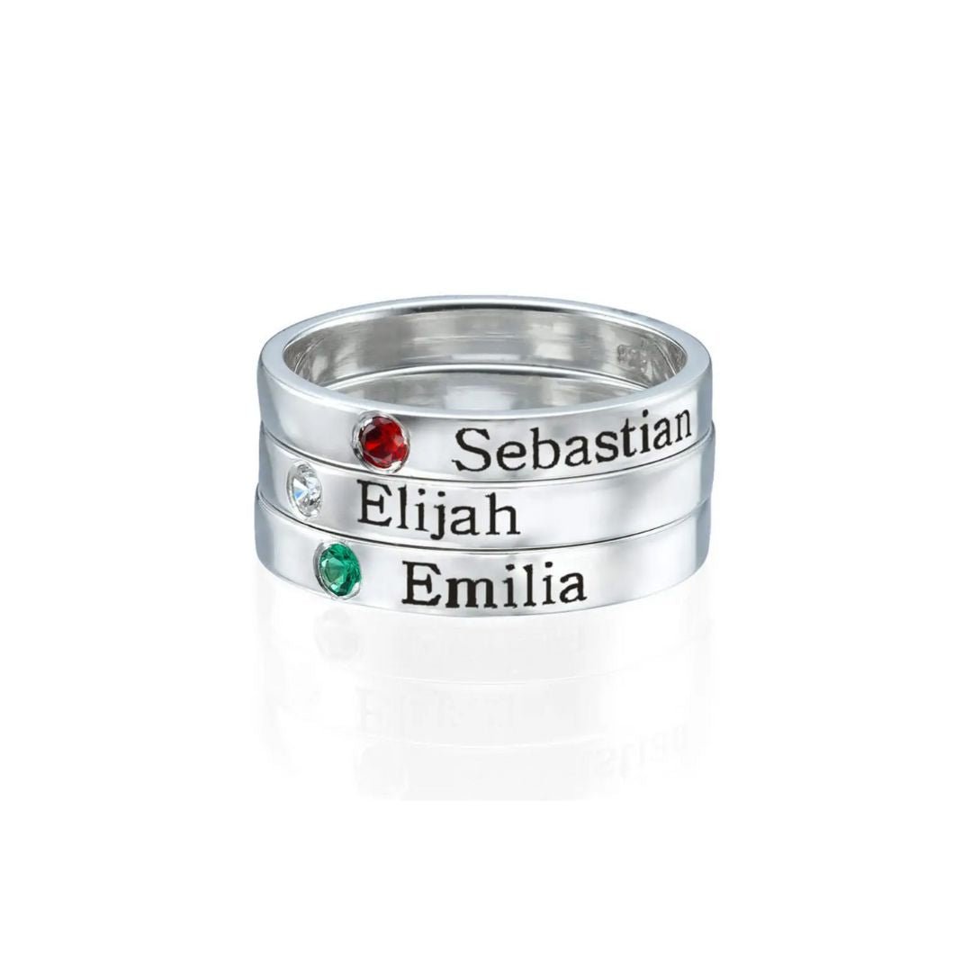 Tres Anillos apilables con nombres y piedras de nacimiento exhiben "Sebastian" (rojo), "Elijah" (transparente) y "Emilia" (verde); cada uno con su nombre grabado y piedra correspondiente.