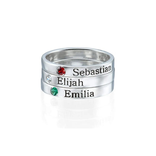 Tres Anillos apilables con nombres y piedras de nacimiento exhiben "Sebastian" (rojo), "Elijah" (transparente) y "Emilia" (verde); cada uno con su nombre grabado y piedra correspondiente.