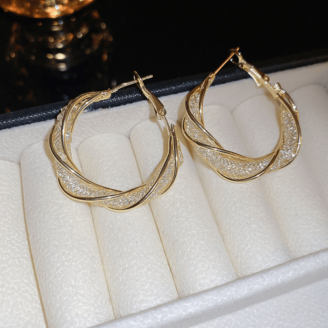 Aretes colgantes en espiral, hechos de acero inoxidable dorado con detalles brillantes, se muestran sobre una bandeja para joyas color crema con borde negro.