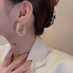 Una mujer con el pelo oscuro recogido en un moño, sujeto con una horquilla, lleva una americana color crema y aretes colgantes en espiral. Su mano cuidada con uñas minimalistas se alza cerca de su cuello.
