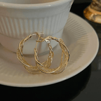 Los Aretes colgantes en espiral, fabricados en acero inoxidable dorado con intrincados detalles retorcidos, se presentan en un platillo blanco delante de una taza blanca acanalada.